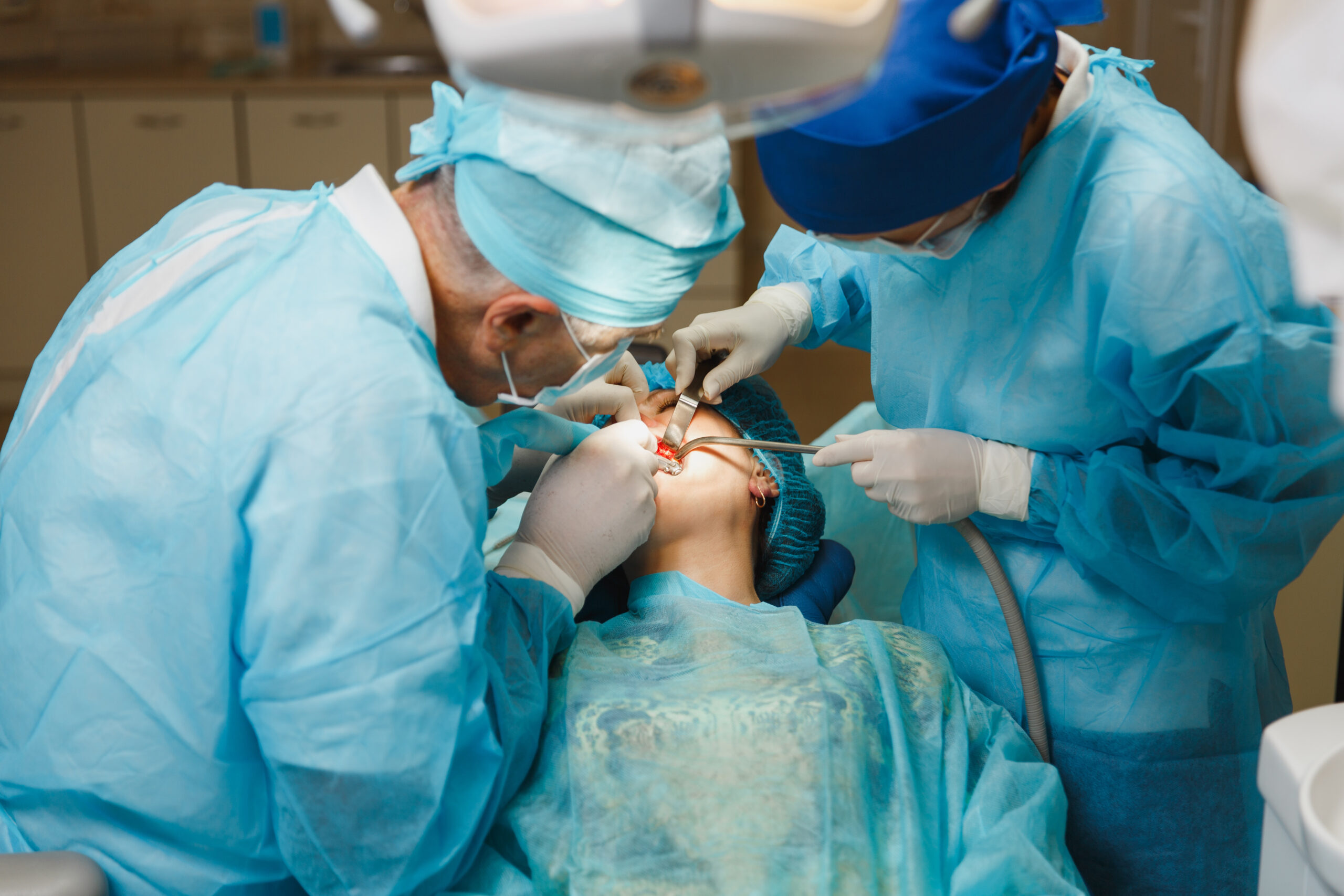 Male old professional dentist surgeon in uniform and female assistant helps to performing operation install dental implant teeth of woman patient in clinic light office with modern tools equipment.